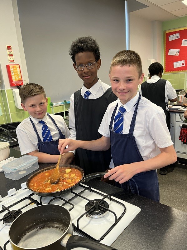 Three students cooking food in a pan over a hob.